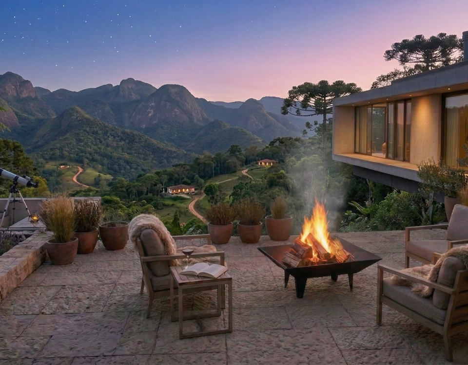 632bd9e3 e5a9 4e68 9114 de77e7fc652d Pátio externo com poltronas ao redor de uma fogueira, vasos de plantas e vista para as montanhas ao pôr do sol, ao lado de uma casa contemporânea de vidro.