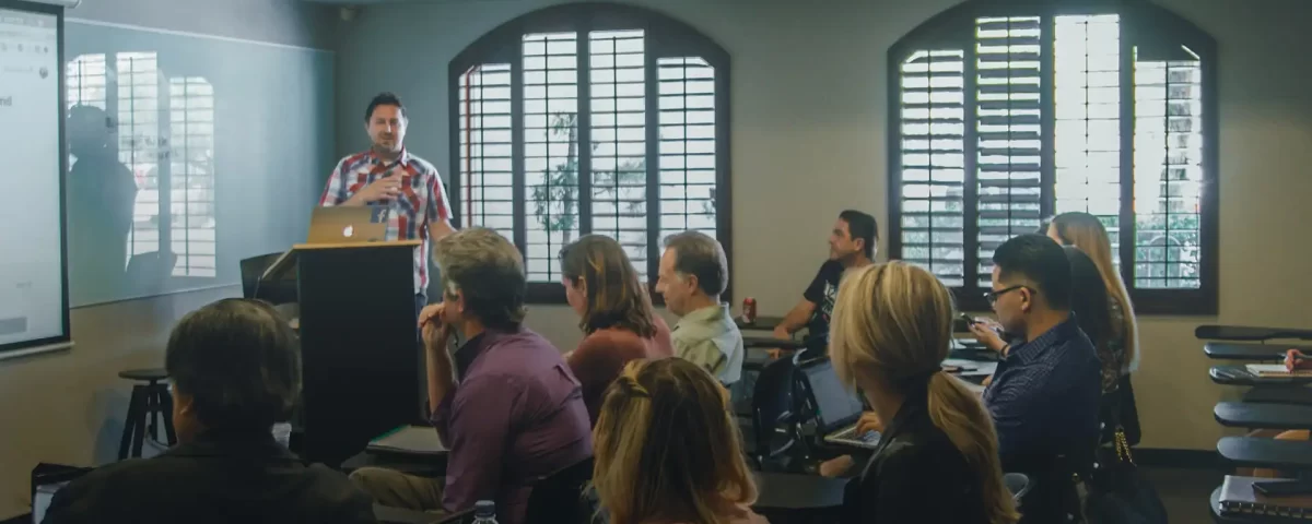 Palestrante em pé ao lado de um púlpito, apresentando-se a uma plateia em sala de conferências, com janelas grandes ao fundo.