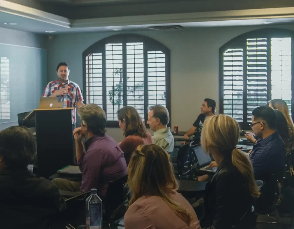 Palestrante em pé ao lado de um púlpito, apresentando-se a uma plateia em sala de conferências, com janelas grandes ao fundo.