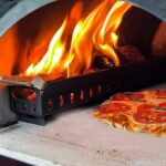 Pizza assando em forno a lenha com chamas visíveis.