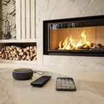 MF Evo Fire 03 1 Sala de estar com lareira de mármore embutida, pilha de lenha ao lado e alto-falante inteligente, controle remoto e smartphone sobre o piso.