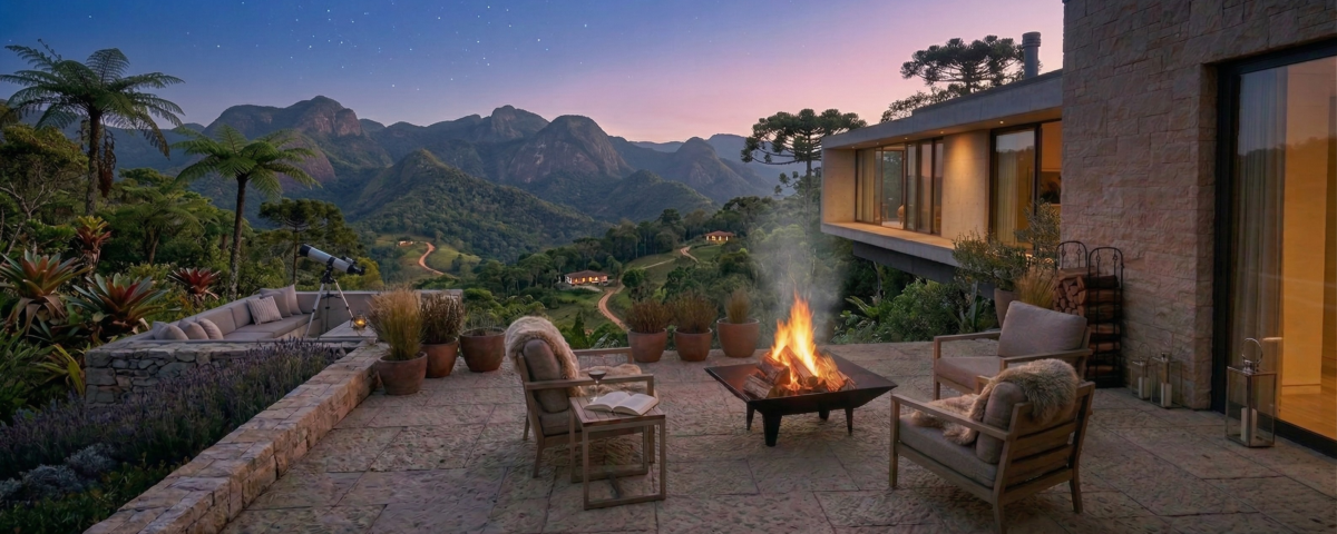 Pátio externo com poltronas ao redor de uma fogueira, vasos de plantas e vista para as montanhas ao pôr do sol, ao lado de uma casa contemporânea de vidro.