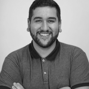 Homem jovem com barba e cabelo curto, usando camisa polo cinza, braços cruzados, sorrindo para a câmera.