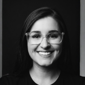 Mulher sorrindo em retrato preto e branco, usando óculos transparentes e camiseta preta.