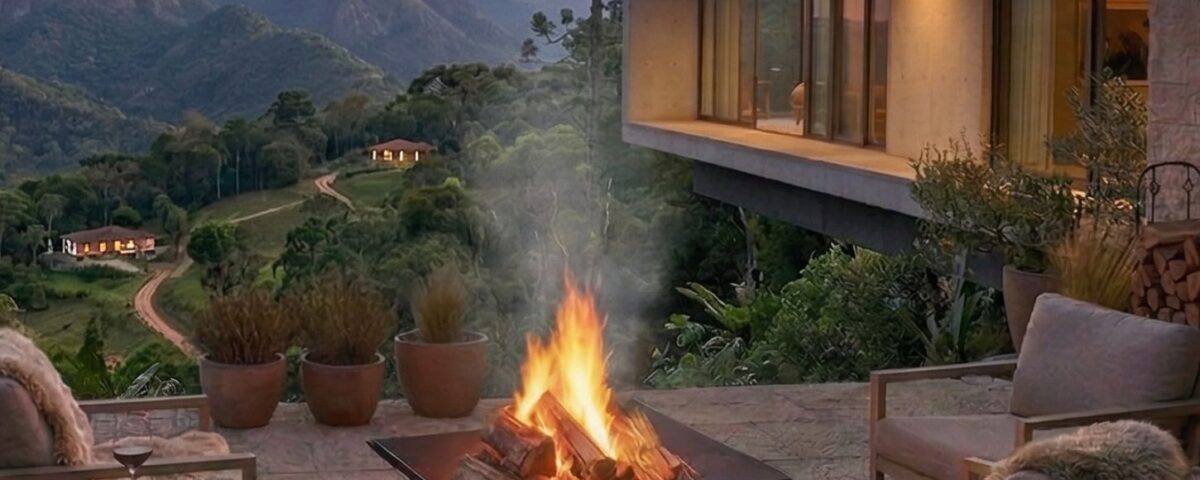 Terraço ao ar livre com lareira acesa, cadeiras aconchegantes e vista de montanhas ao pôr do sol.