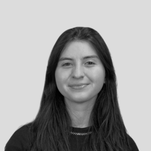Mulher jovem com cabelo longo e escuro, vestindo blusa preta, sorrindo levemente, com fundo cinza claro.